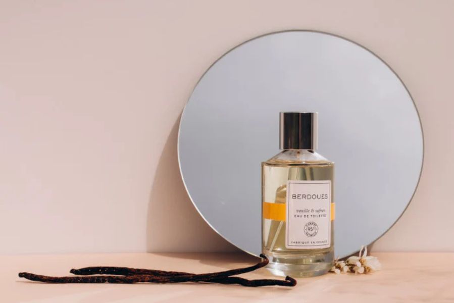 Flacon d’Eau de Toilette Berdoues Vanille & Safran posé devant un miroir rond, accompagné de gousses de vanille, symbole d’élégance gourmande.