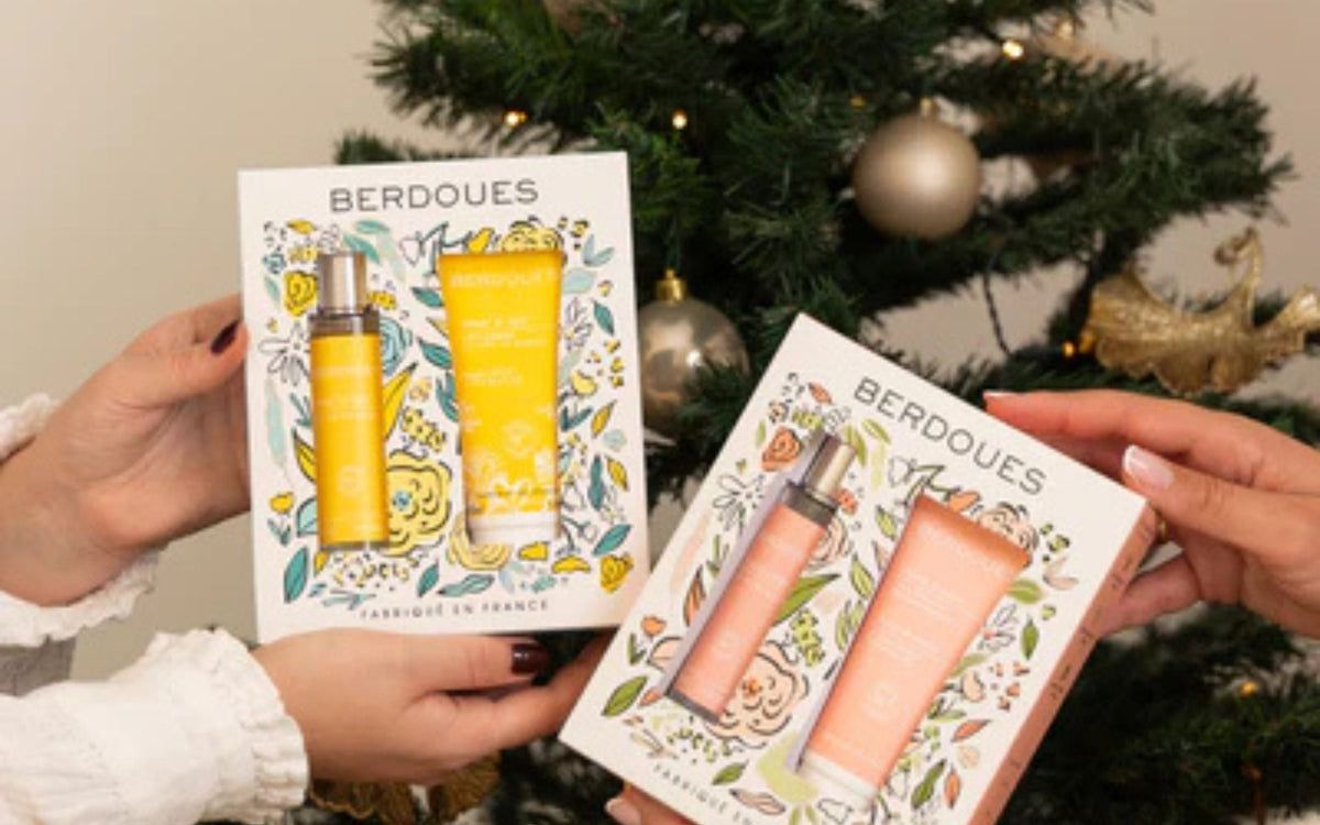 Deux coffrets parfum et soin Berdoues tenus en main devant un sapin de Noël.
